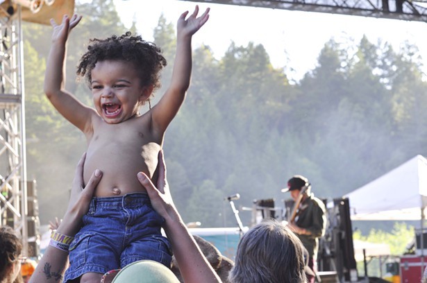 Indubious bassist/lyricist Spencer “Skip Wicked” Burton plays in the background at Summer Arts and Music Festival in Benbow. His newest, biggest, smallest fan approves. - ©TRAVIS TURNER