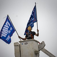 A masked parade participant waves campaign flags from a crane bucket above a truck parked at the end of the caravan in McKinleyville.
