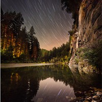 The South Fork Eel River, giant redwood trees and wheeling stars overhead making streaks over the course of a 21:02-minute exposure. The reddish light in the trees is from dwellings across the river. Redway Beach, Redway, California. Oct. 6, 2023.